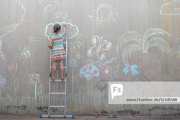 Mädchen steht auf einer Leiter und zeichnet bunte Bilder mit Kreide auf einer Betonwand.