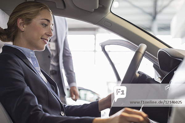 Lächelnde Frau beim Stöbern im neuen Auto, auf dem Fahrersitz im Autohaus sitzend