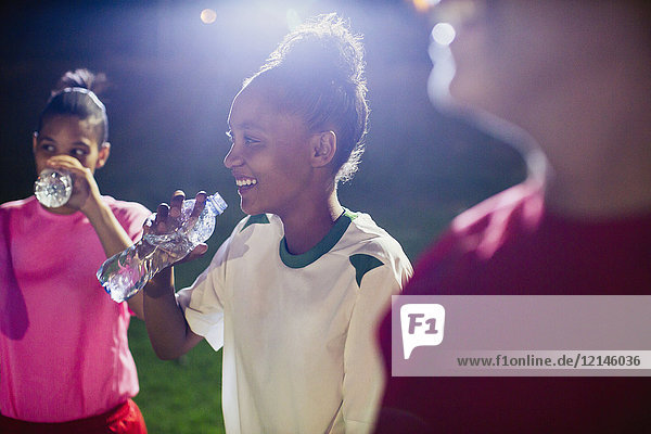 Smiling young female soccer players resting  drinking from water bottles on field at night