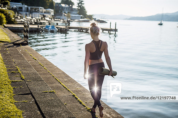Rückansicht der Frau in Sportbekleidung mit Yogamatte am Seeufer