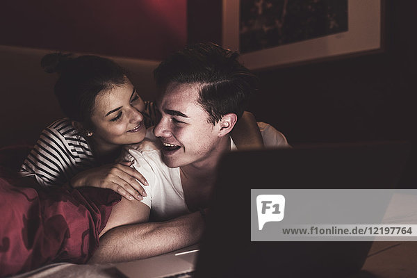 Happy couple lying in bed at home with laptop