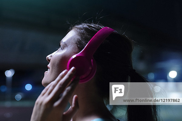 Happy young woman with pink headphones listening to music in modern urban setting at night