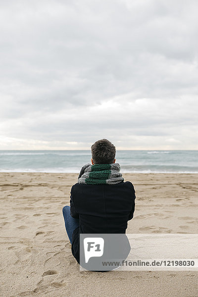 Rückansicht des Mannes  der im Winter am Strand sitzt und in die Ferne schaut.