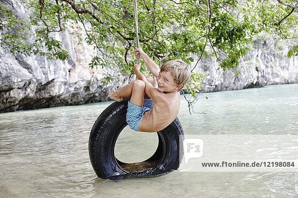 Thailand  Ko Yao Noi  Phang Nga Bay  glücklicher Junge  der auf Reifen über dem Meer schwingt.