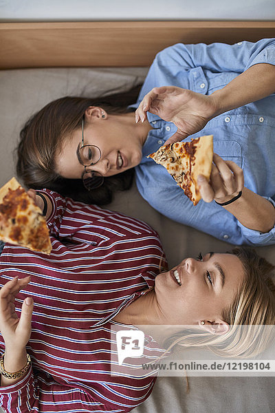 Two young women lying down eating pizza together