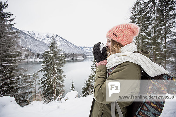 Junge Frau mit heißem Getränk stehend in alpiner Winterlandschaft mit See