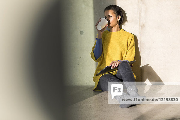 Junge Frau im gelben Pullover sitzt auf dem Boden und trinkt Kaffee zum Mitnehmen.