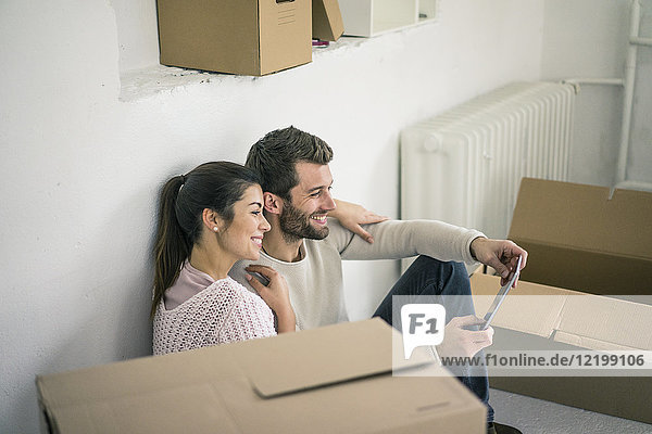 Couple sitting in new home surrounded by cardboard boxes looking at tablet