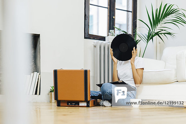 Young woman sitting on grounf listening music from record player  hiding behind vinyl