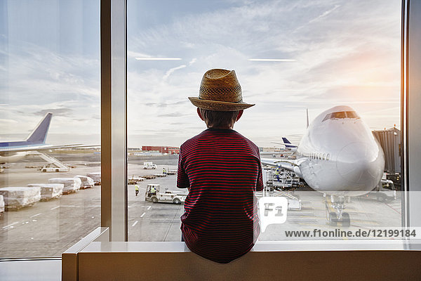 Junge mit Strohhut und Blick durchs Fenster zum Flugzeug auf dem Vorfeld