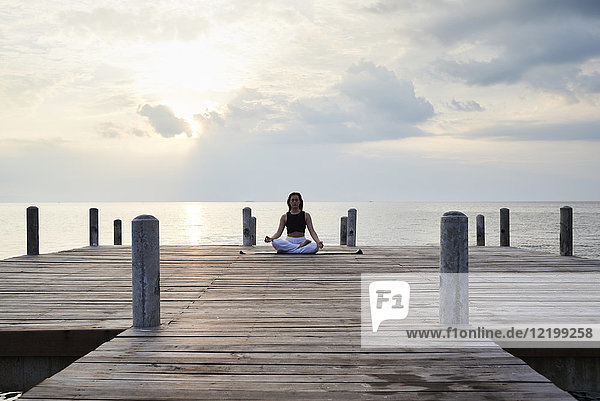 Junge Frau praktiziert Yoga an einem Steg am Meer bei Sonnenuntergang