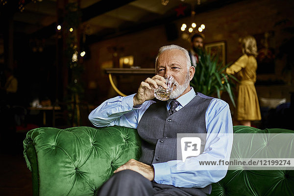 Porträt eines eleganten älteren Mannes  der auf einer Couch in einer Bar sitzt und aus einem Becher trinkt.