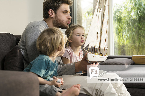 Vater und Kinder blasen in die Segel eines Spielzeugmodellschiffes auf der Couch zu Hause.