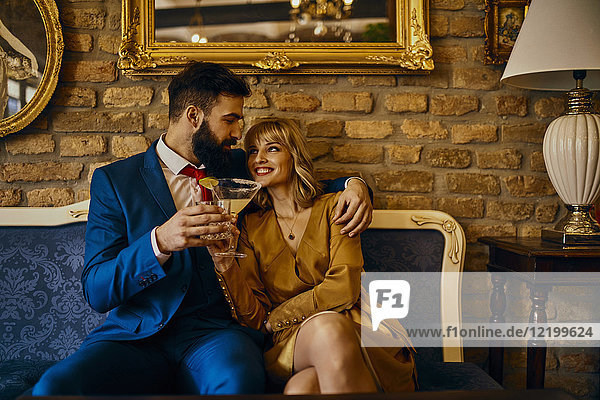 Happy elegant couple with drinks sitting on couch embracing