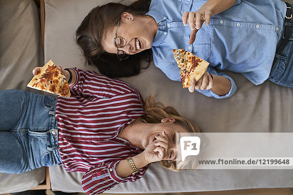 Two laughing young women lying down eating pizza together