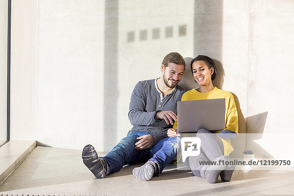 Porträt eines lachenden jungen Paares auf dem Boden sitzend mit Laptop