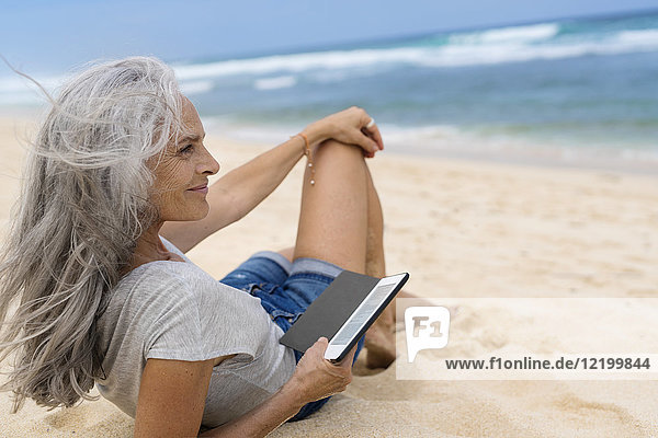 Schöne lächelnde Seniorin am Strand liegend,  mit E-Book