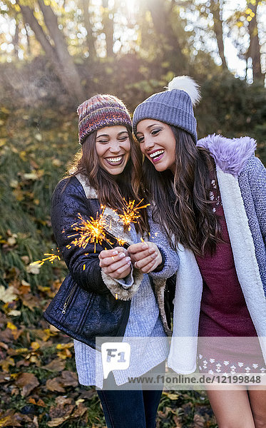 Zwei fröhliche Frauen halten Wunderkerzen in einem herbstlichen Wald.