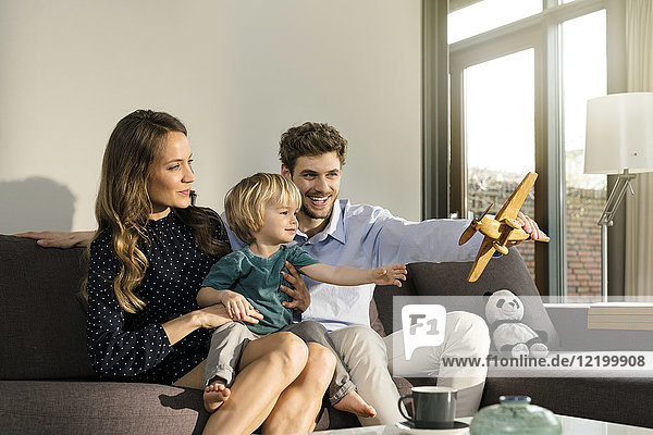Happy parents and son playing with wooden toy plane on sofa at home
