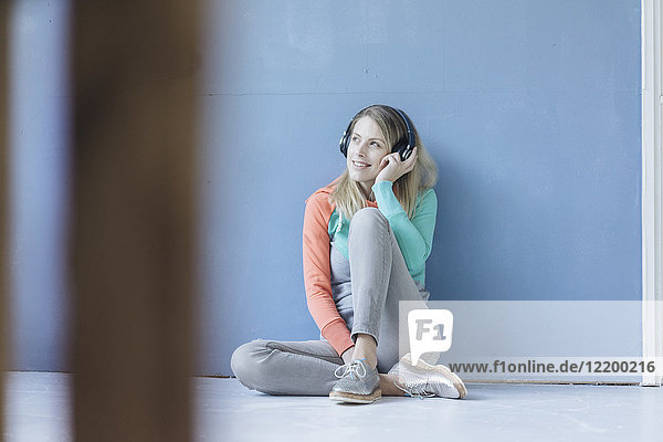 Porträt einer lächelnden Frau,  die auf dem Boden vor einer blauen Wand sitzt und Musik mit kabellosen Kopfhörern hört.