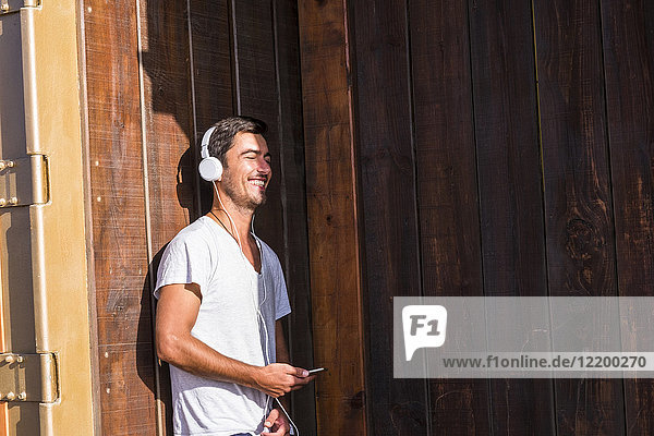 Happy young man with cell phone and headphones leaning against wooden wall