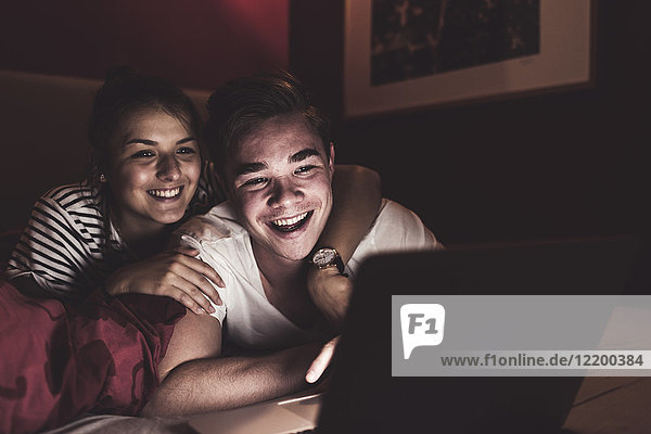 Happy couple lying in bed at home looking at laptop