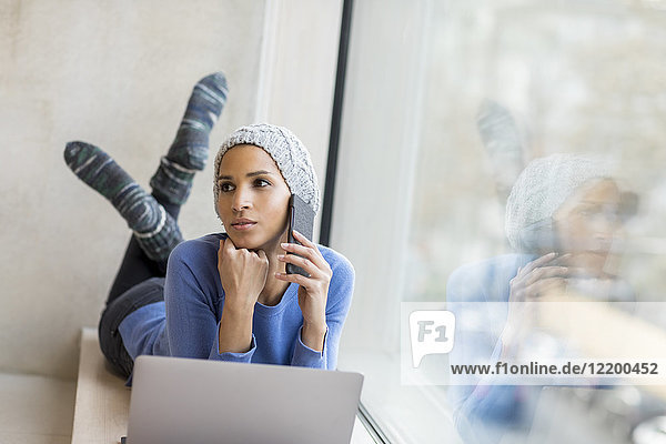 Porträt einer jungen Frau am Telefon liegend mit Laptop auf Fensterbank
