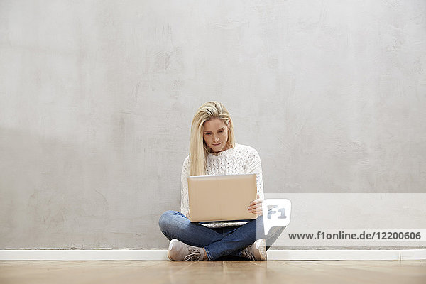 Blond woman sitting on the floor in front of grey wall using laptop