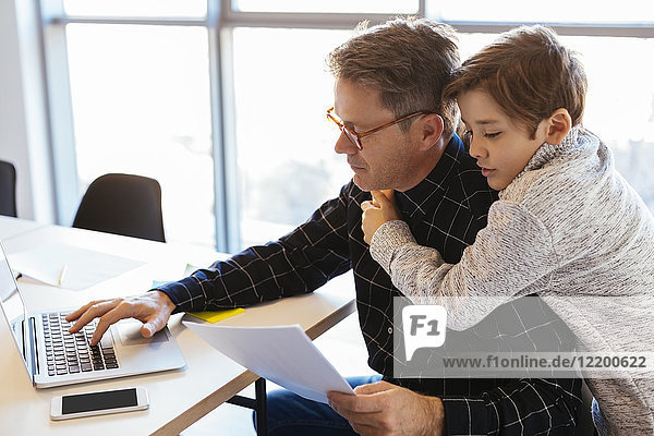 Businessman using laptop at desk in office with son embracing him