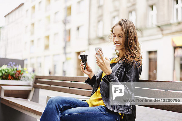 Porträt einer glücklichen Frau  die auf einer Bank mit Eistüte sitzt und auf ihr Handy schaut.