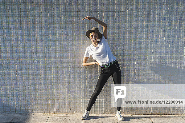 Smiling woman wearing hat standing in front of tiled wall doing stretching exercises