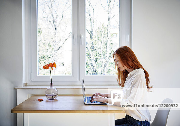 Rothaarige Frau sitzt am Tisch vor dem Fenster mit Laptop