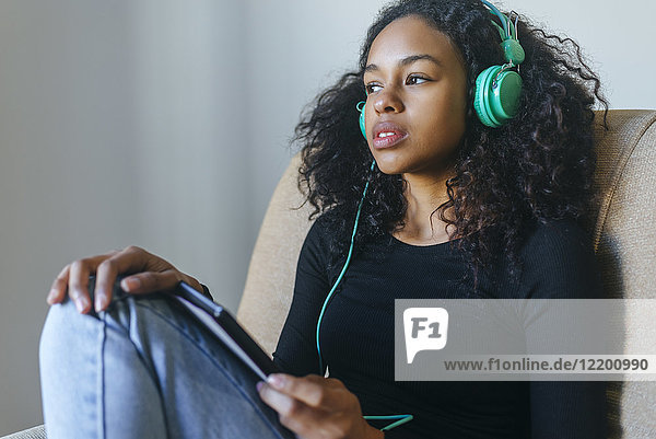Portrait einer jungen Frau  die im Sessel sitzt und Musik mit Kopfhörer und Tablett hört.