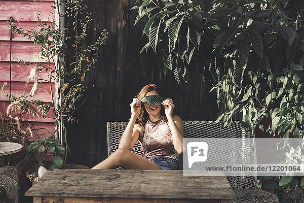 Junge Frau sitzt auf einer Bank im Freien und bedeckt die Augen mit einem Blatt.