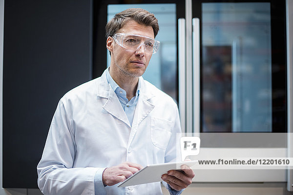 Porträt eines Mannes mit Laborkittel und Schutzbrille  der die Tablette an der Maschine hält.