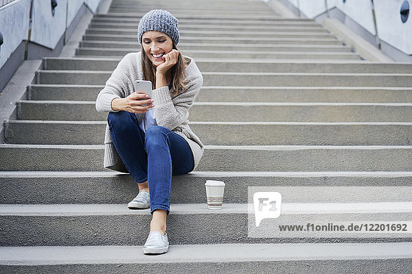 Fröhliche Frau mit Kaffee zum Sitzen auf der Treppe mit Blick auf das Handy