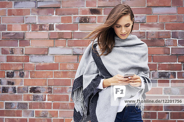Smiling young woman looking at cell phone in front of brick wall