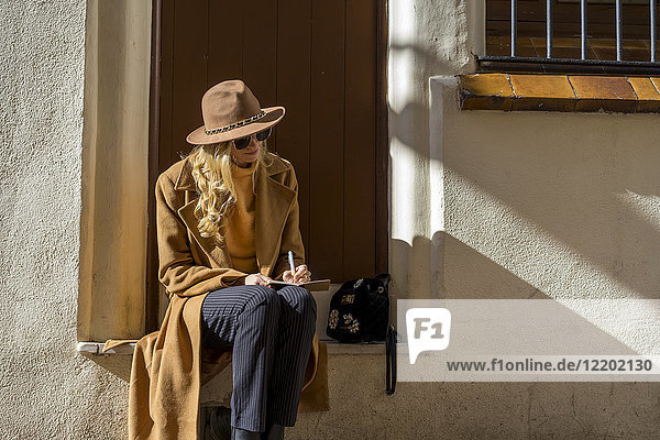 Modische junge Frau sitzt am Hauseingang und schreibt in ein Notizbuch.