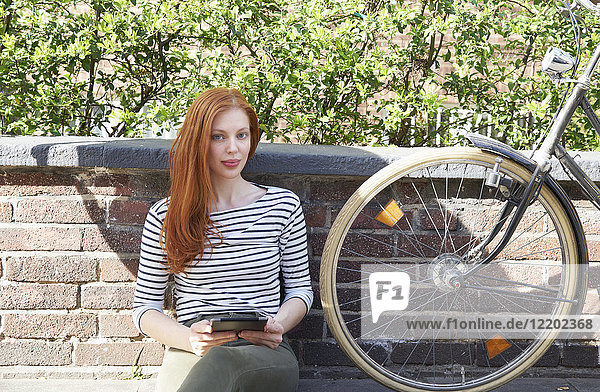 Porträt einer rothaarigen Frau mit Tablette und Fahrrad auf einer Wand sitzend
