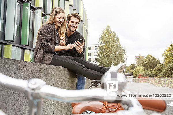 Happy couple with bicycles sitting on a wall looking at cell phone