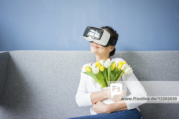 Lächelnde Frau sitzt auf der Couch und trägt eine VR-Brille mit Tulpen.