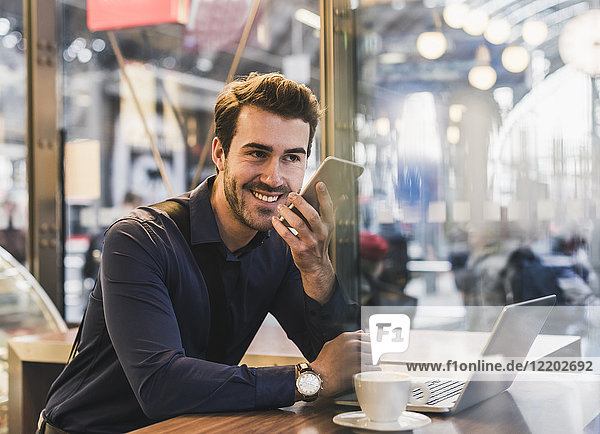 Lächelnder junger Geschäftsmann im Café am Bahnhof mit Handy und Laptop