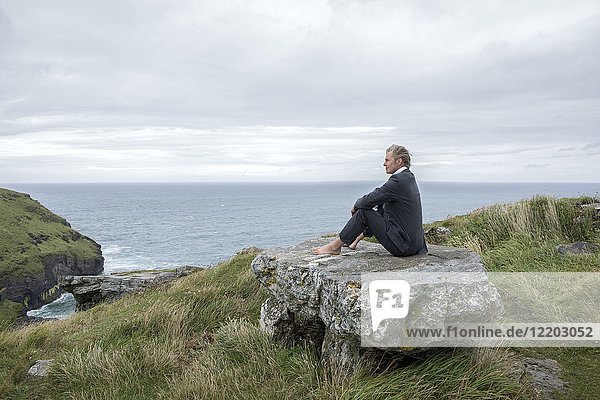 UK  Cornwall  Tintagel  businessman sitting on rock at the coast looking at view
