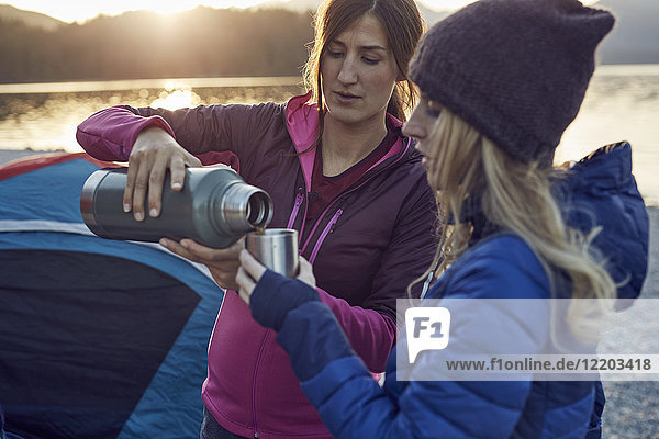 Two women with hot drink camping at lakeshore