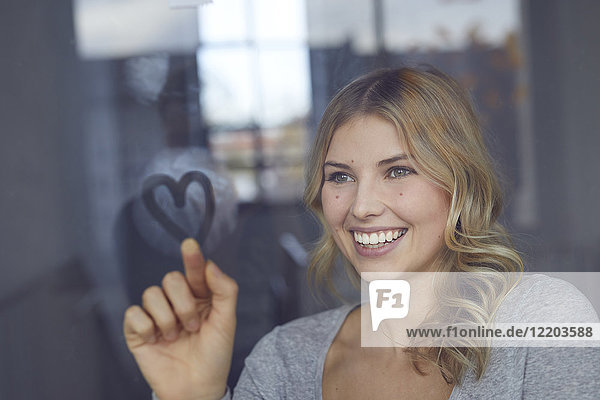 Porträt einer fröhlichen blonden Frau  die ein Herz auf einer Fensterscheibe zeichnet