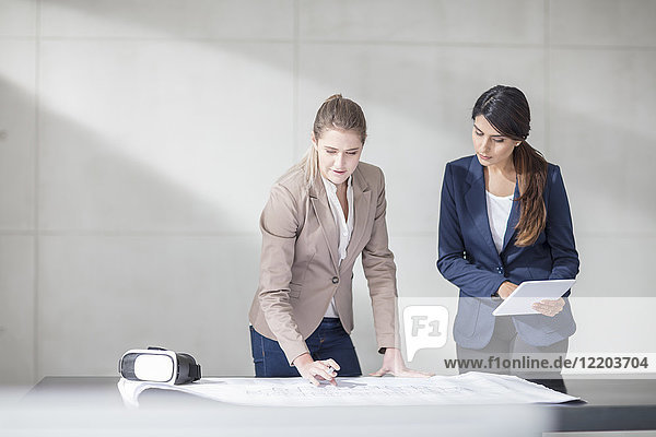 Zwei junge Frauen mit Tablette und VR-Brille bei der Arbeit an der Blaupause im Büro
