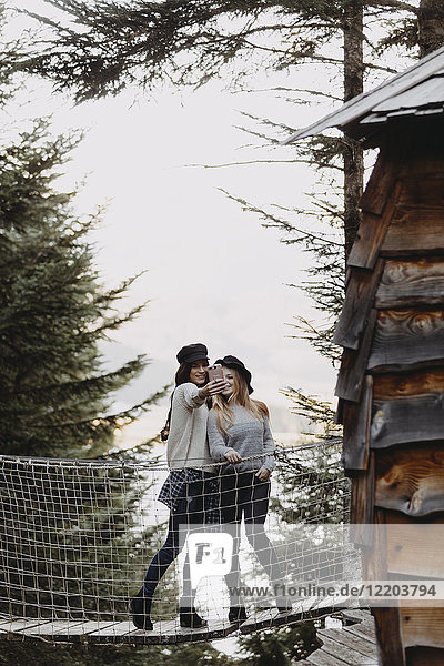 Zwei junge Frauen auf einer Hängebrücke bei einem Baumhaus im Wald mit einem Selfie
