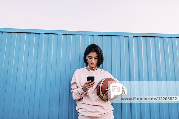 Junge Frau mit Basketball  Smartphone und Kopfhörer am Container