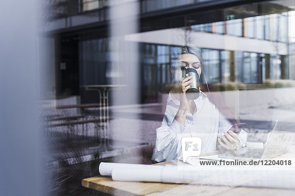 Junge Frau mit Laptop  Handy und Unterlagen beim Kaffeetrinken hinter der Fensterscheibe
