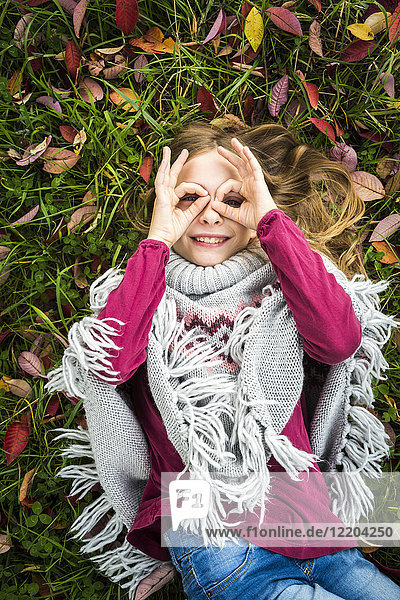 Portrait of smiling girl lying on autumnal meadow looking through her fingers formed like spectacles